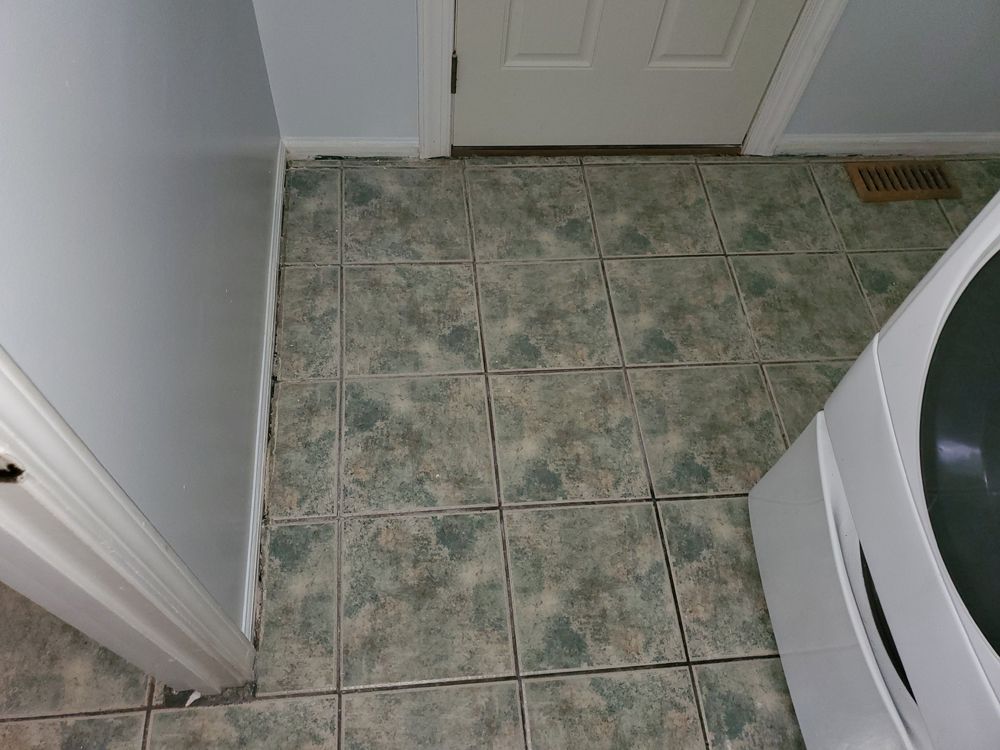 Tiled floor with a washing machine on the right, a door in the background, and a gray wall on the left.