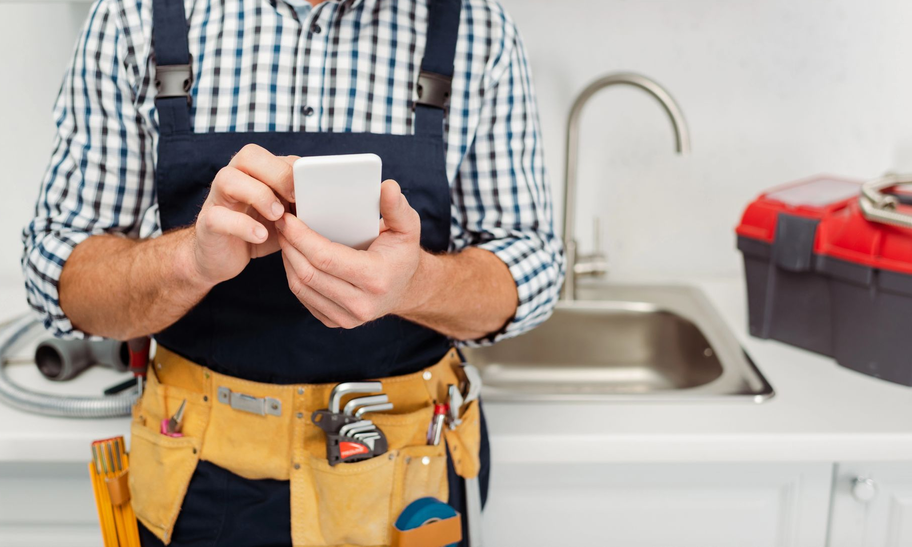 Plumber in overalls holding a phone near a sink and toolbox.
