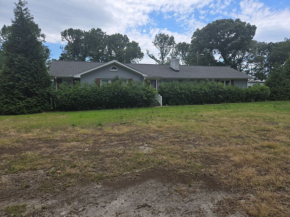 Gray house with a dark roof, surrounded by bushes, set on a grassy lawn under a cloudy sky.