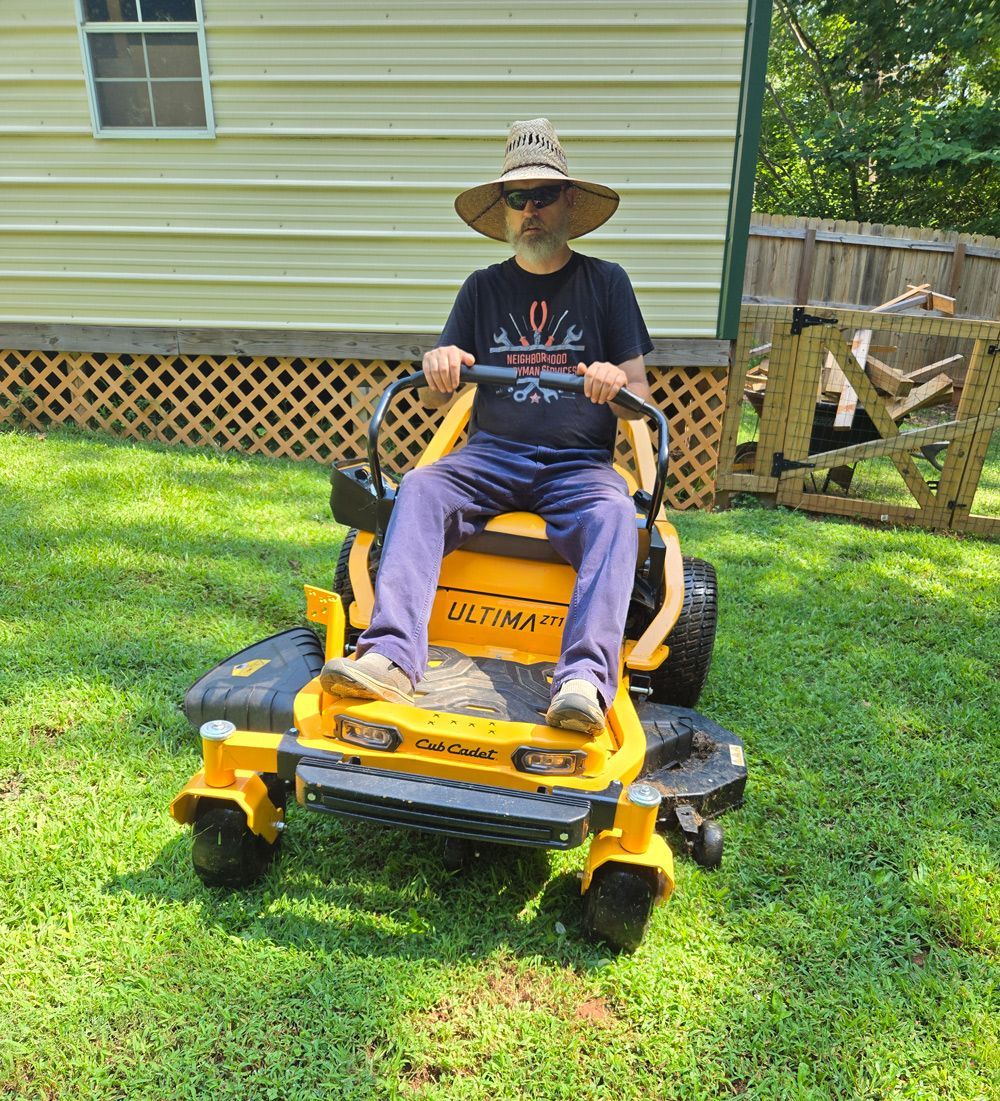 Man wearing a straw hat on a yellow riding lawnmower in a backyard.