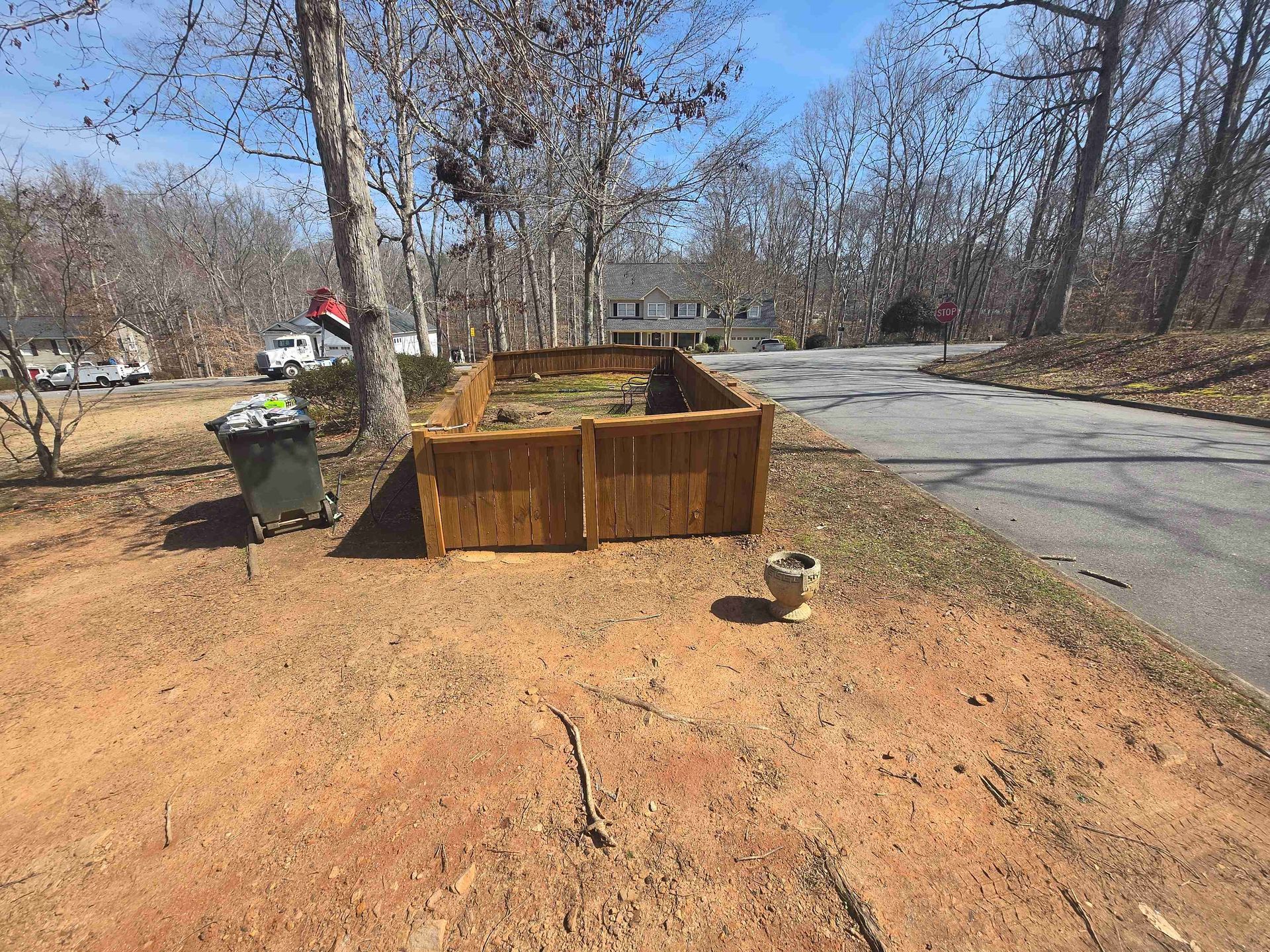 A wooden raised garden bed sits on a dirt lot next to a driveway, with a trash bin and bare trees in the background.