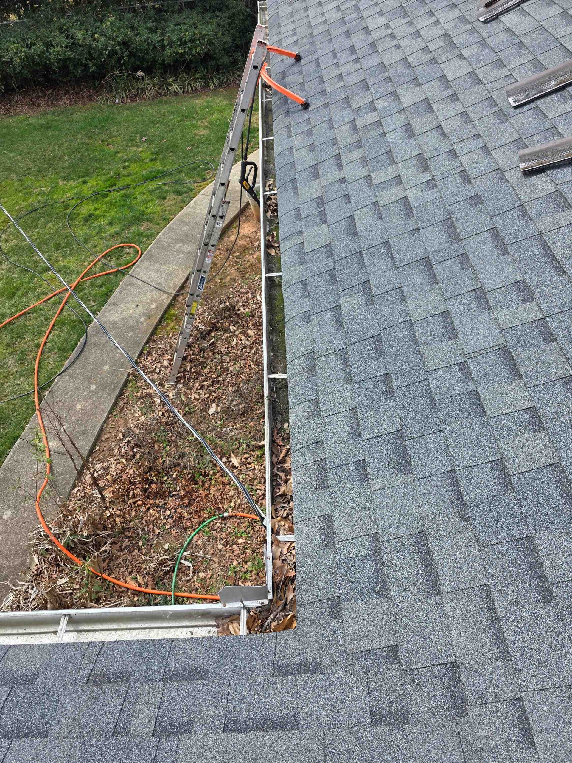A metal ladder leans against a house with grey shingles, positioned next to a gutter filled with dead leaves and debris.