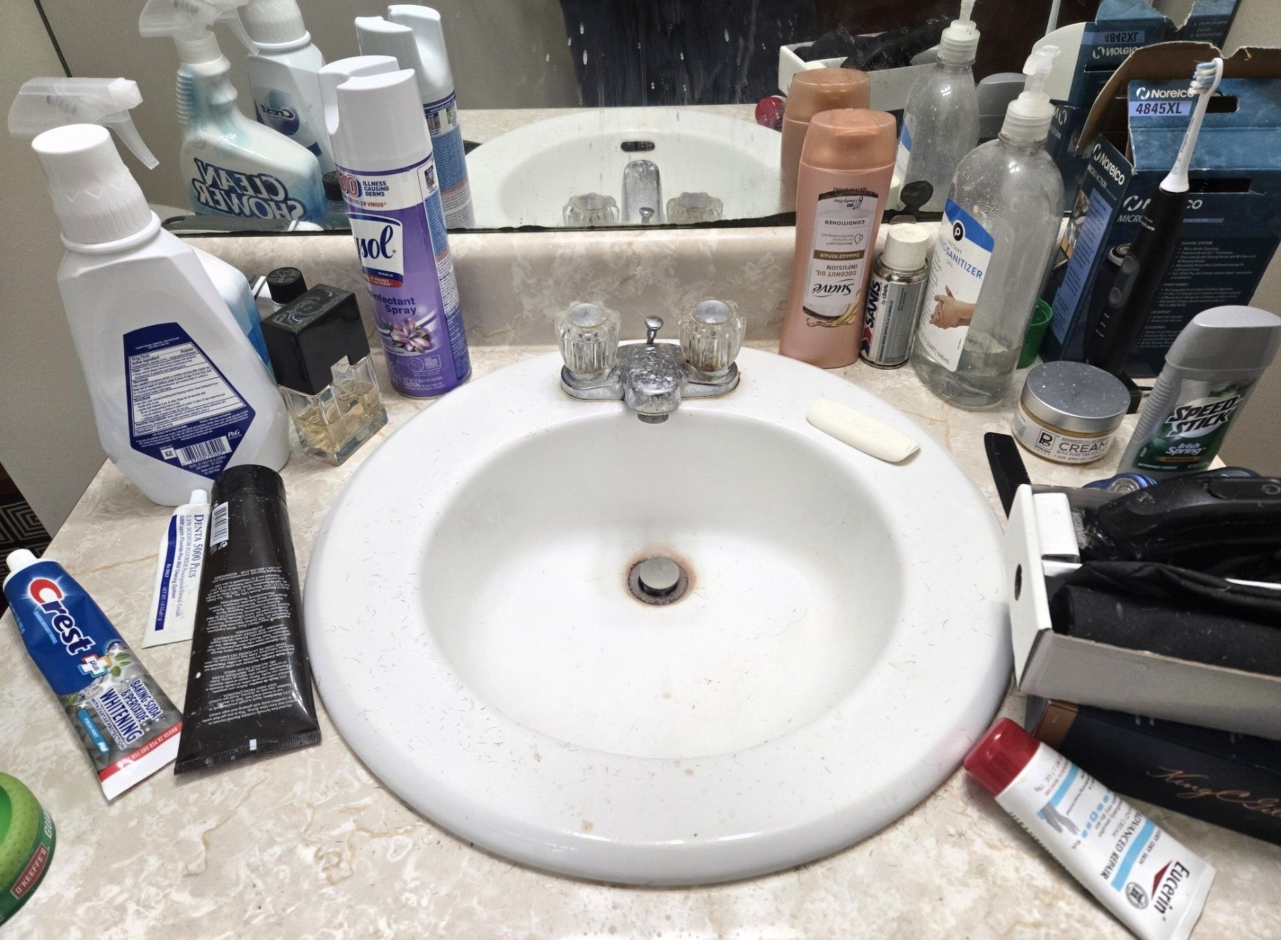 A cluttered bathroom sink counter filled with various toiletries, cleaning bottles, and personal care products.