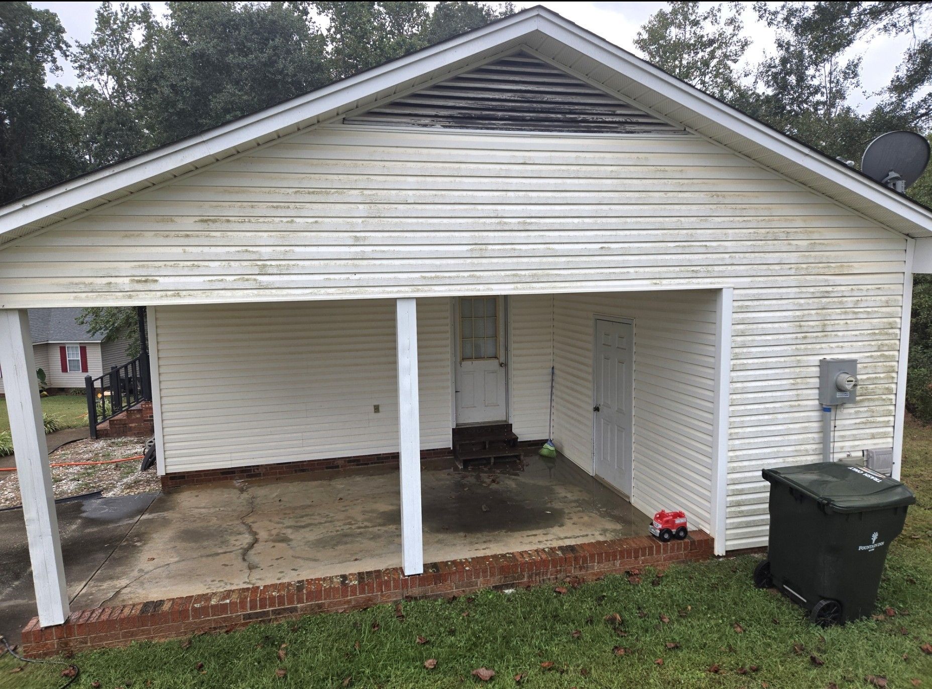 A weathered white house exterior with a covered porch, brick base, and a dark garbage bin on the grass.