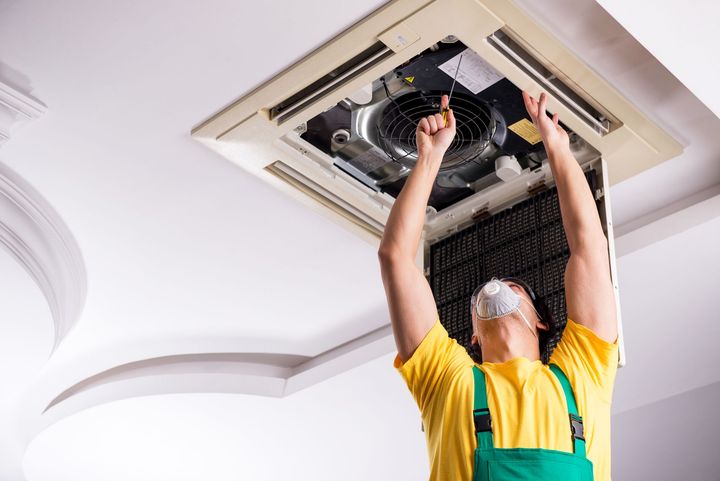 A man in a yellow shirt and green overalls is working on an air conditioner.