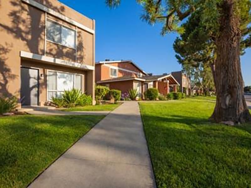 Apartment complex with a walkway, green lawns, and trees under a clear blue sky.