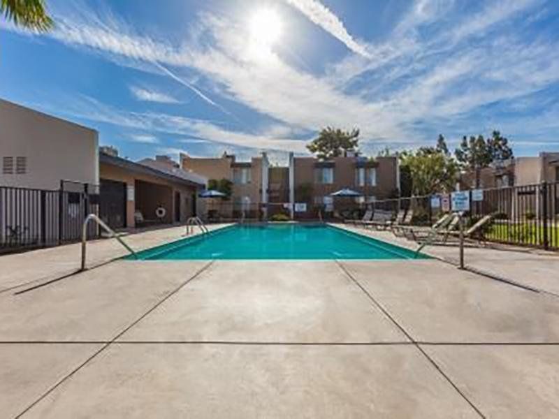 Swimming pool surrounded by concrete, fence, and buildings under a sunny blue sky.