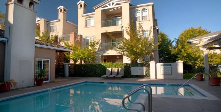 Swimming pool in front of beige apartment building with trees, and clear blue sky.