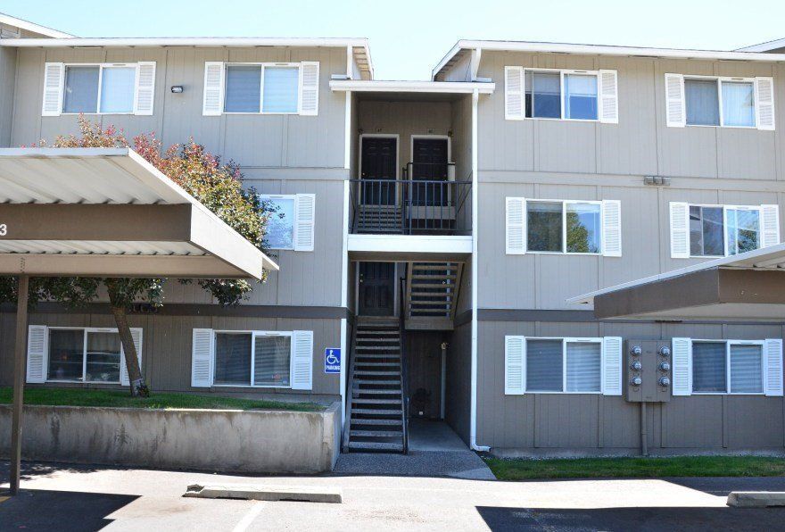 Apartment building with gray siding, white shutters, and a central staircase. Includes a handicap parking sign and carports.