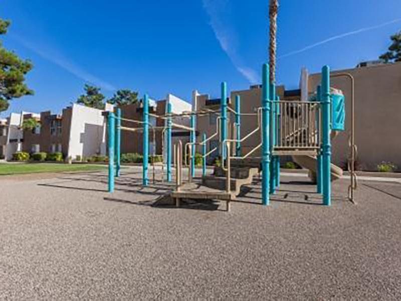 Playground with blue equipment on a pebbled surface, in front of buildings, under a blue sky.