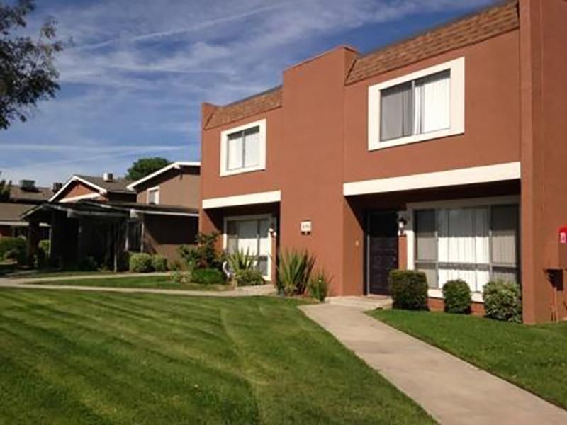 Brown two-story apartment building with white window trim on a sunny day with green grass and a sidewalk.