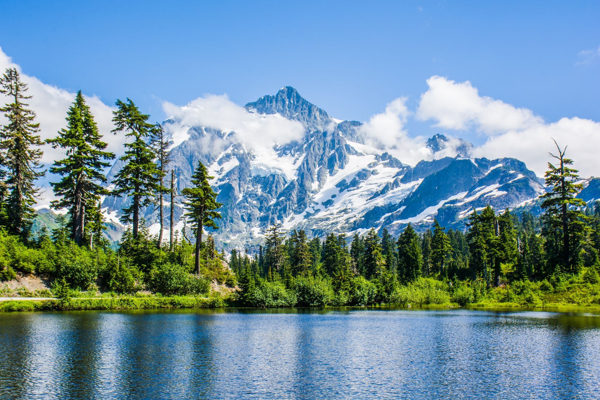 Snow-capped mountains rise behind a lake and evergreen trees under a blue sky with fluffy white clouds.