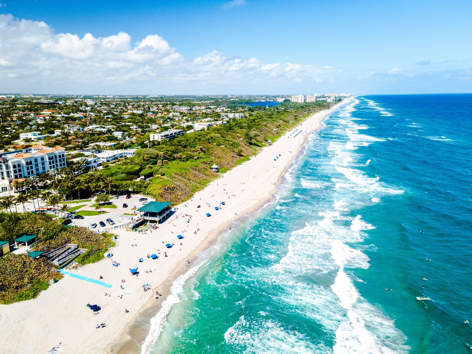 Aerial view of a sunny beach with turquoise water, white sand, and a line of buildings and trees.