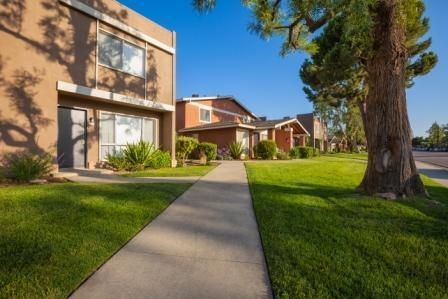 Sidewalk leading to apartments with brown facades, green grass, and blue sky.
