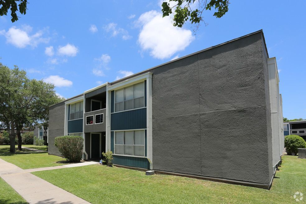 Two-story apartment building with stucco exterior and teal accents, set in a grassy yard under a blue sky.