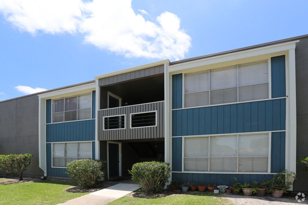 Apartment building exterior, two stories, blue siding, white-trimmed windows, gray accents, small bushes, blue sky.