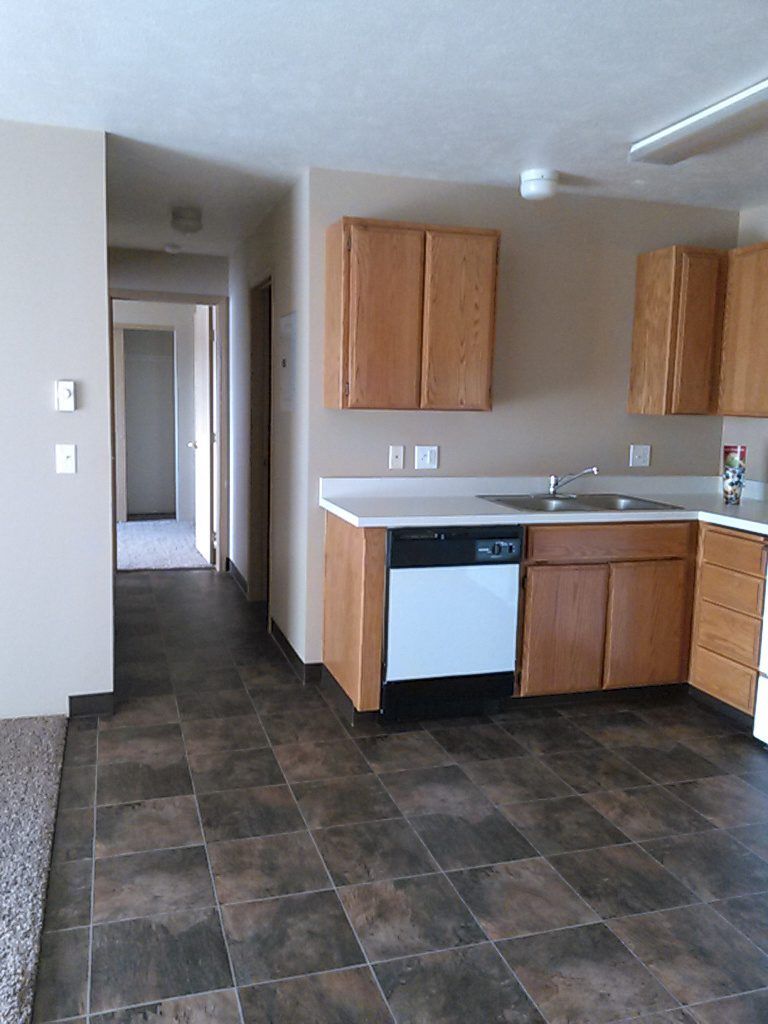 Kitchen with wood cabinets, white appliances, and brown tile flooring, leading into a hallway.