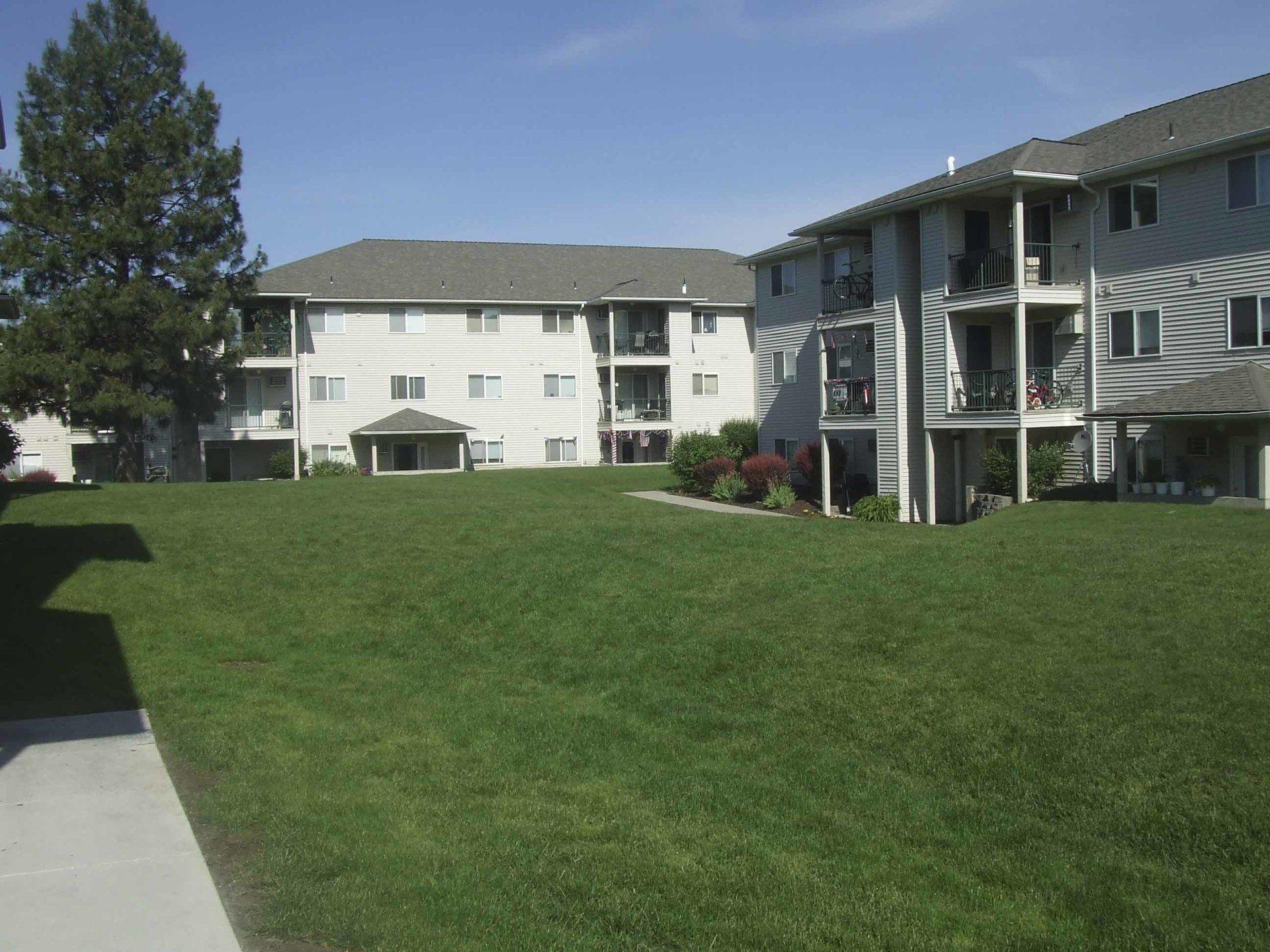 Apartment buildings with multiple floors and balconies, surrounded by green grass on a sunny day.