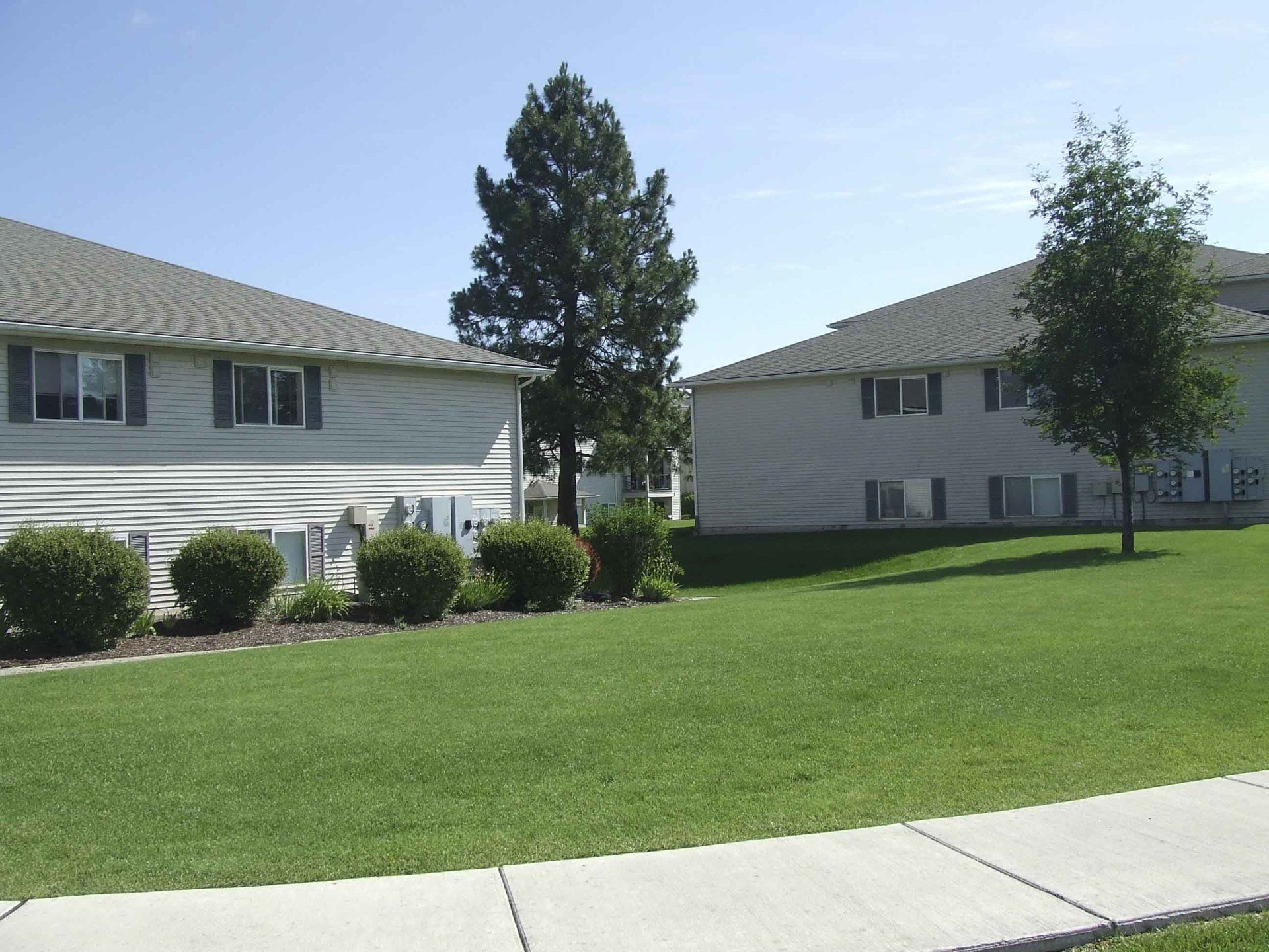 Two-story apartment buildings with light siding and gray roofs, green lawn, bushes, and trees on a sunny day.