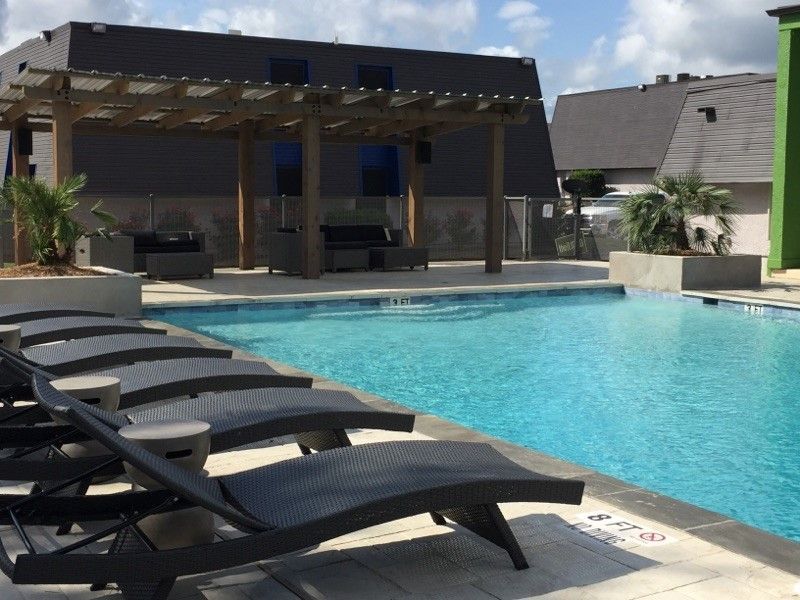 Swimming pool with lounge chairs, pergola, and modern buildings under a sunny sky.