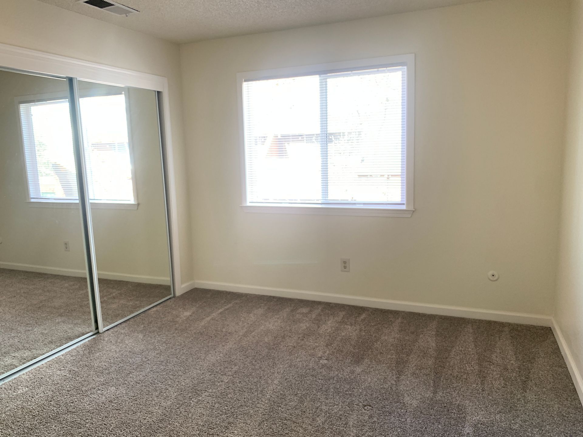 Empty bedroom with mirrored closet doors, window, and brown carpet. Beige walls.