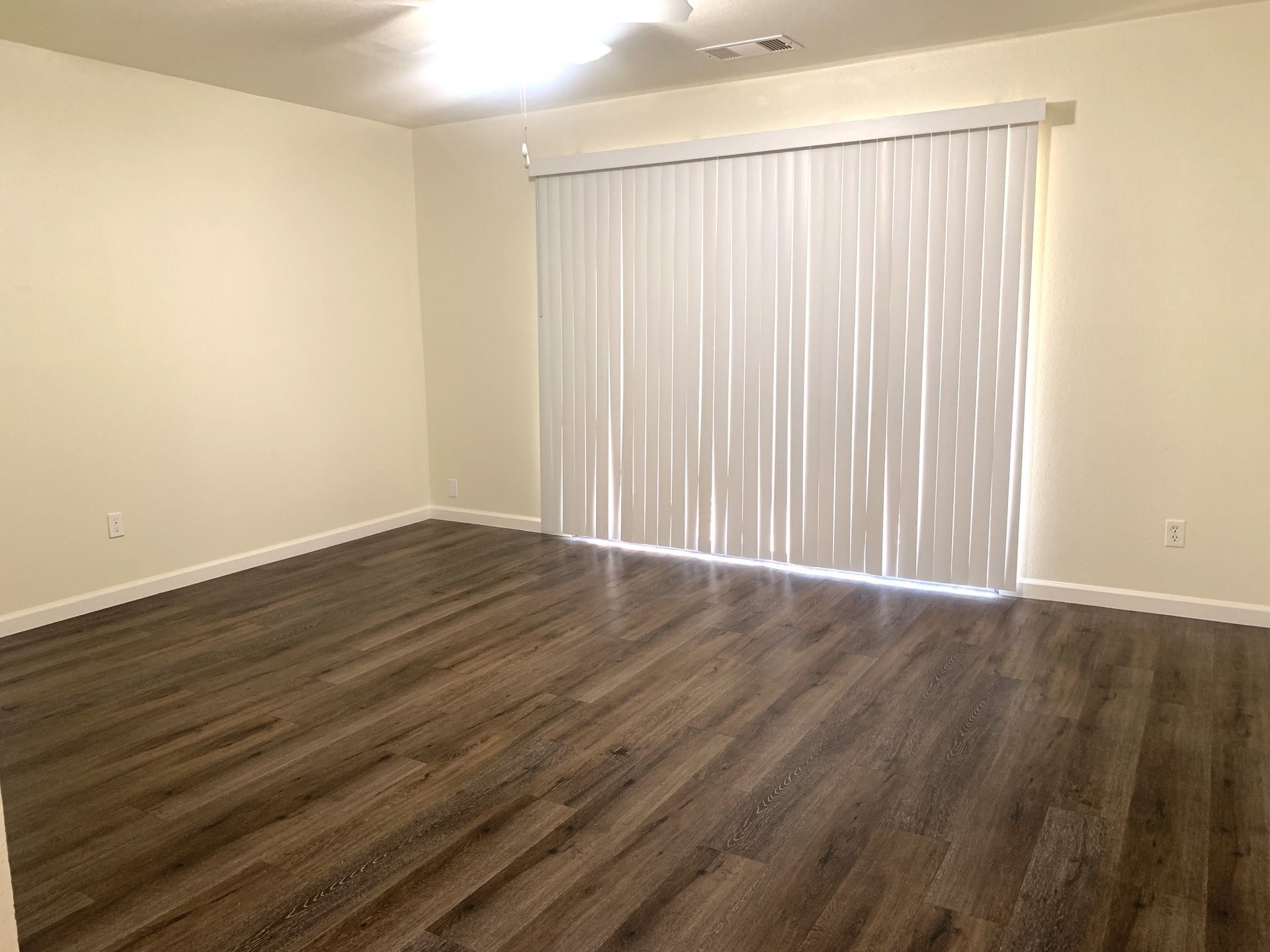 Empty room with wood-look flooring and vertical blinds. Pale walls and bright overhead lighting.