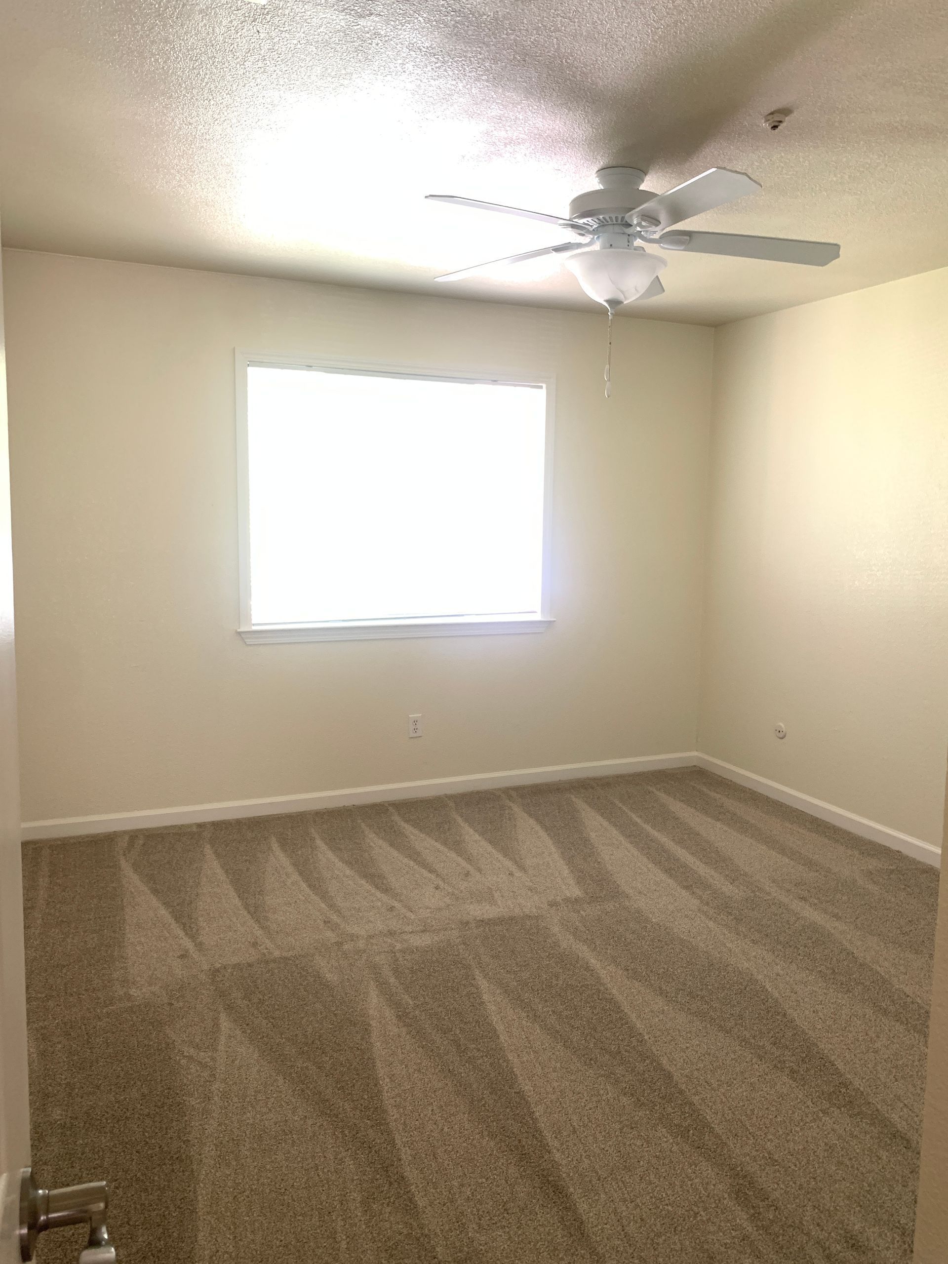 Empty room with beige walls, patterned carpet, window, and ceiling fan.