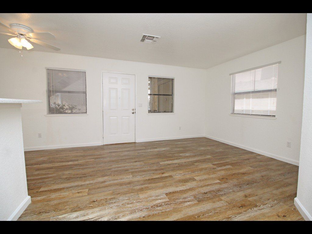 Empty living room with wood-look flooring, white walls, three windows with blinds, and a white door.