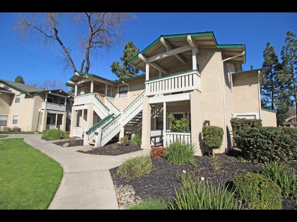 Apartment complex with beige buildings, green trim, and staircases. Path leads to the entrance. Blue sky.