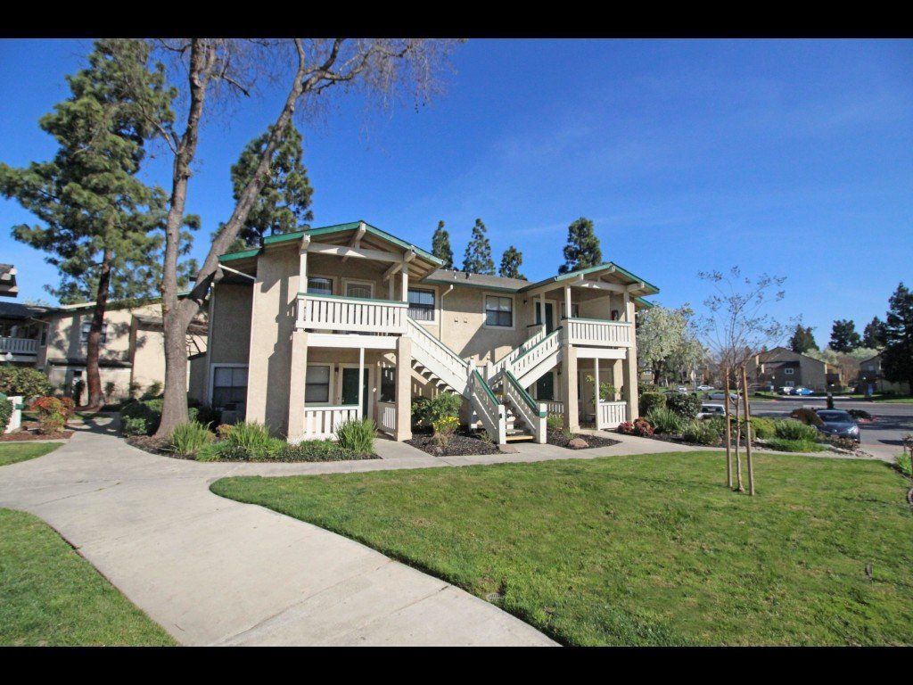 Apartment complex with beige buildings, balconies, green lawn, trees, and blue sky.