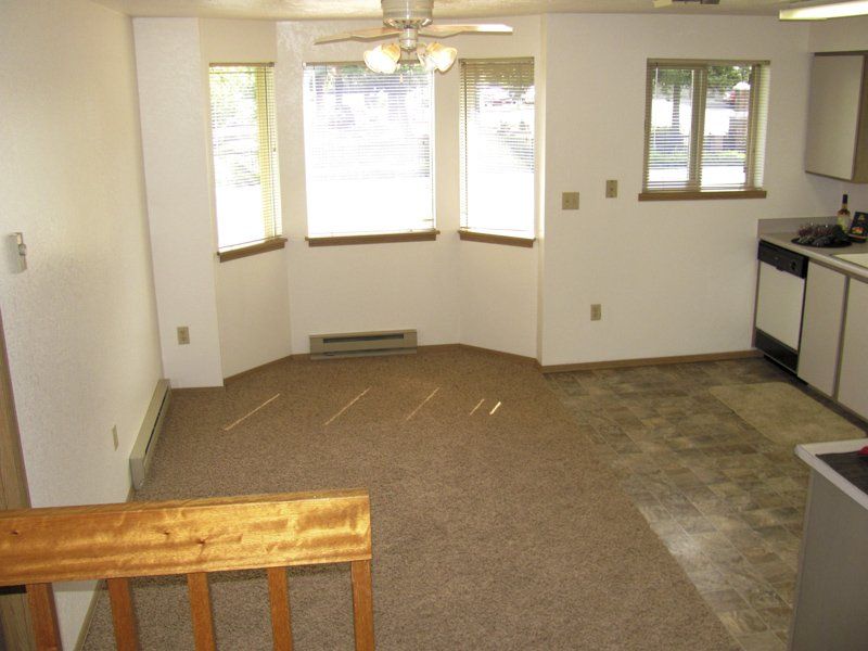 Interior view of a living/dining area with carpet, a bay window, and a small kitchen visible.