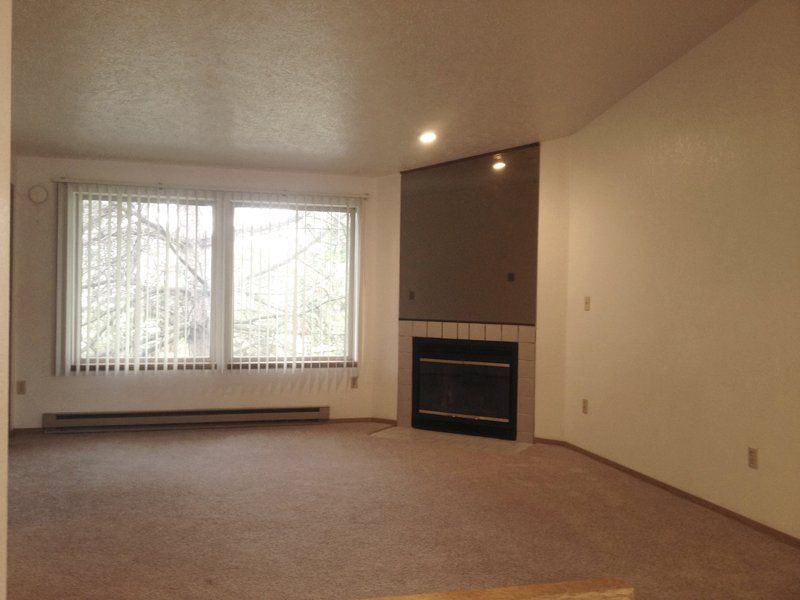 Empty living room with fireplace and large window with blinds. Beige carpet and walls.