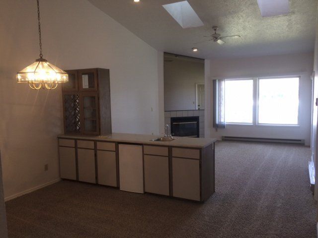Living room with built-in bar, fireplace, and large windows. Brown carpet, beige cabinets, and vaulted ceiling.