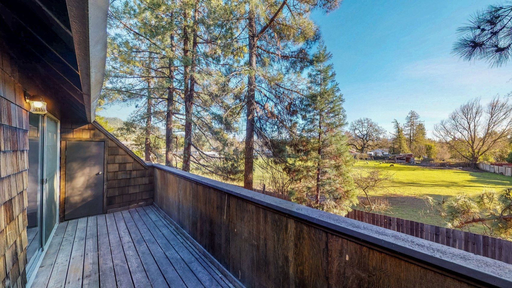 Wooden deck with railing overlooking a sunny, grassy field and trees.