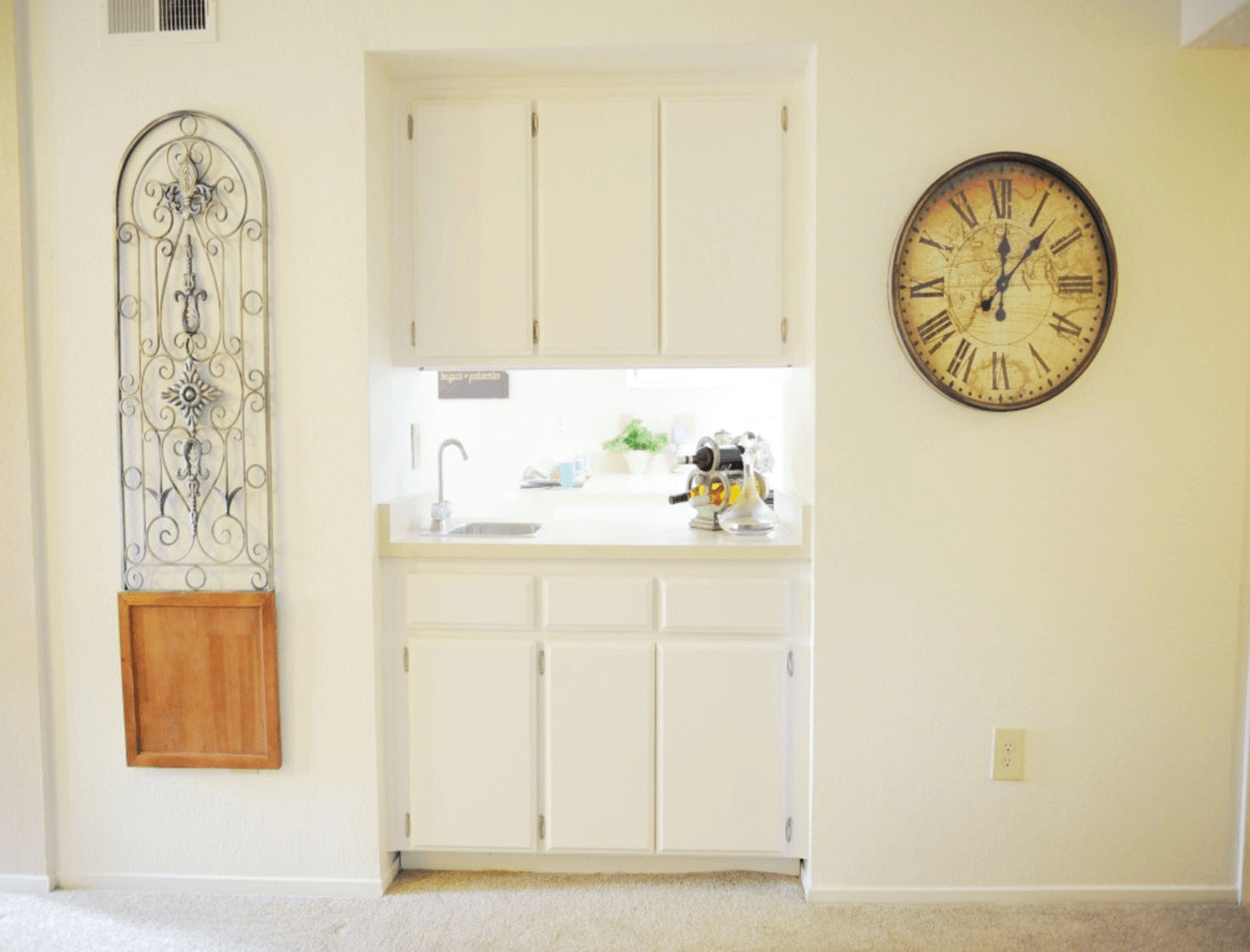 Built-in white cabinets with sink and accessories, a large clock, and a decorative metal piece on a light beige wall.