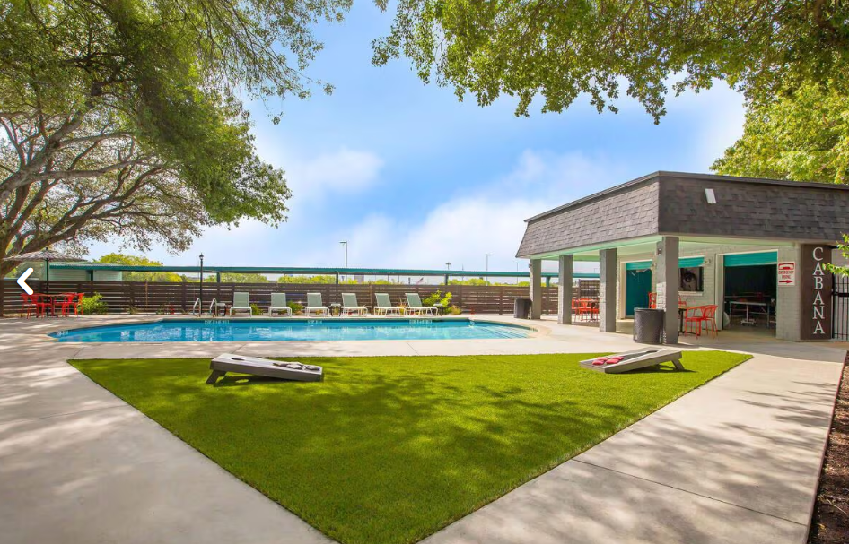 Pool area with blue water, green grass, a covered seating area, and cornhole boards.