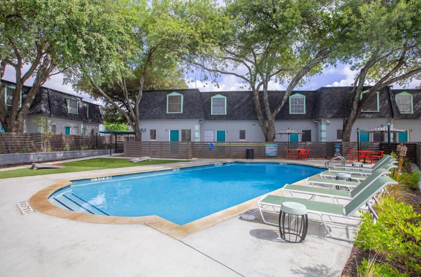 Pool and lounge chairs in front of apartment buildings with turquoise doors and a row of trees.
