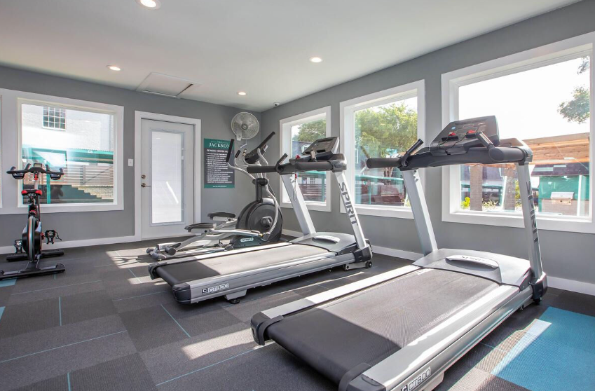 Gym with treadmills, elliptical, and exercise bike near windows and door. Gray walls and flooring.