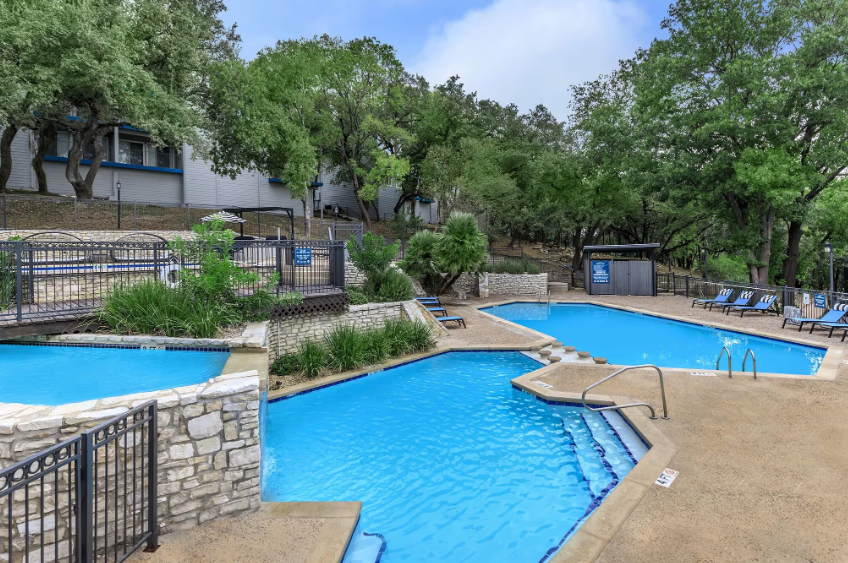 Multiple blue swimming pools surrounded by a stone deck, trees, and buildings on a sunny day.