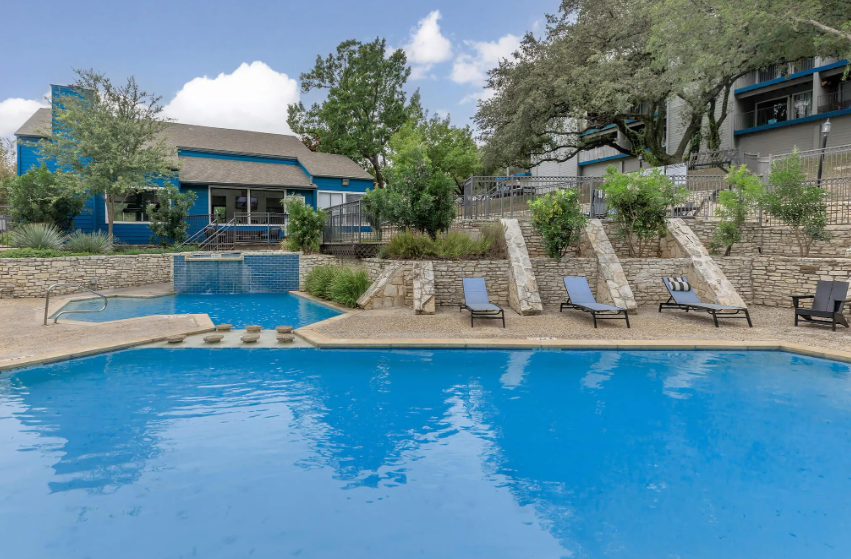 Pool with blue water and lounge chairs near a blue building with trees.