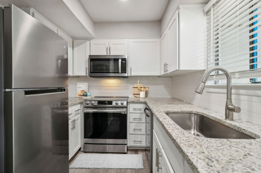 Small, white kitchen with stainless steel appliances, granite countertops, and a window with blinds.