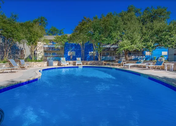 Pool surrounded by lounge chairs, trees, and a blue-painted building under a clear blue sky.