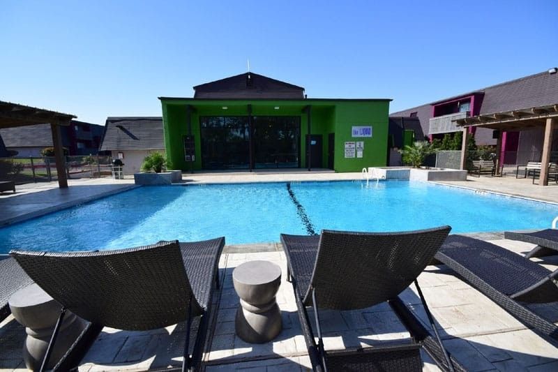 Pool with lounge chairs in front; green building and blue sky in background.