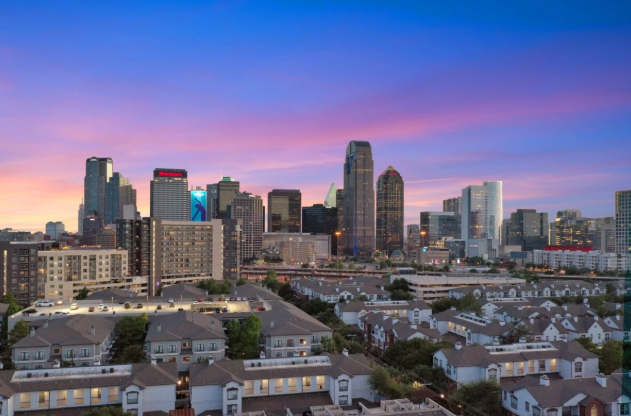 Aerial view of a sunny beach with turquoise water, white sand, and a line of buildings and trees.