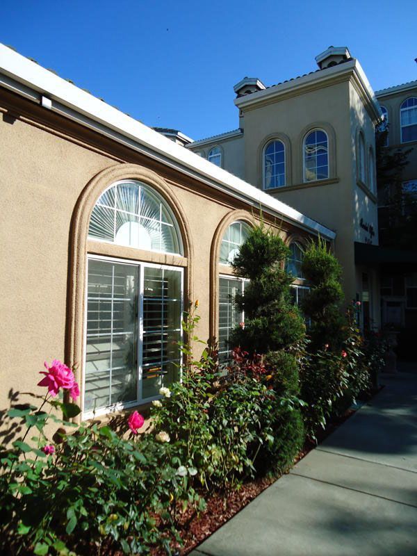 Tan building with arched windows and a garden of flowers and bushes in front.