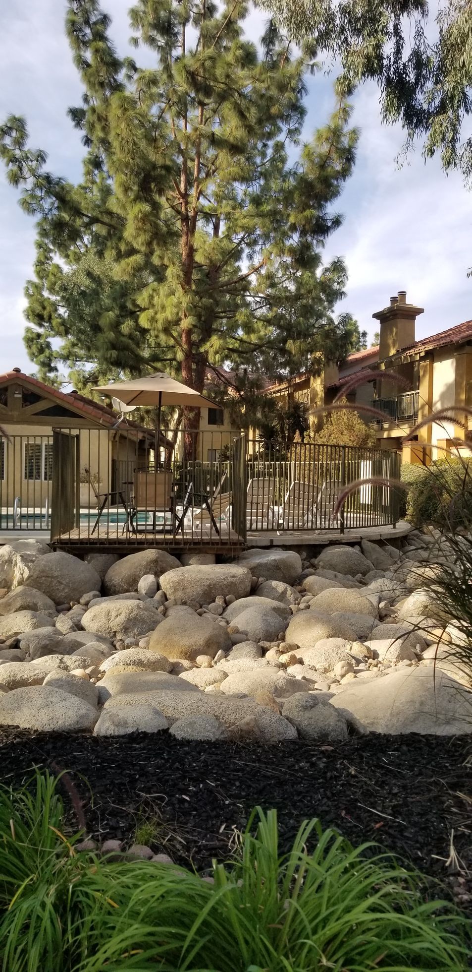 A landscape of large rocks, greenery, and wooden structures, beneath tall trees, on a sunny day.