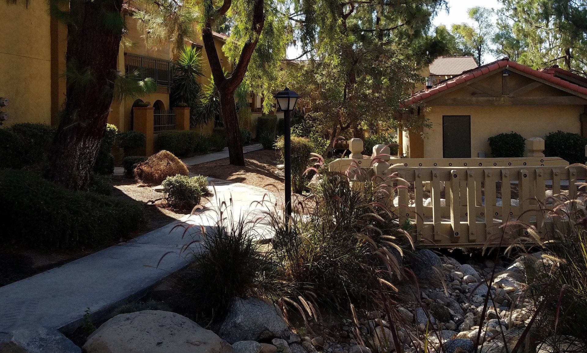 Pathway through a residential area with a bridge and trees. Buildings have tan walls and red tile roofs.