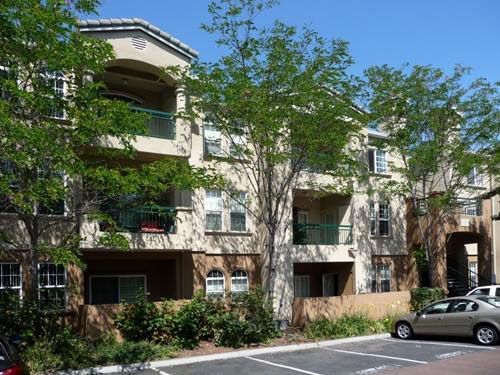 Multi-story beige apartment building with balconies, trees, and parked cars.