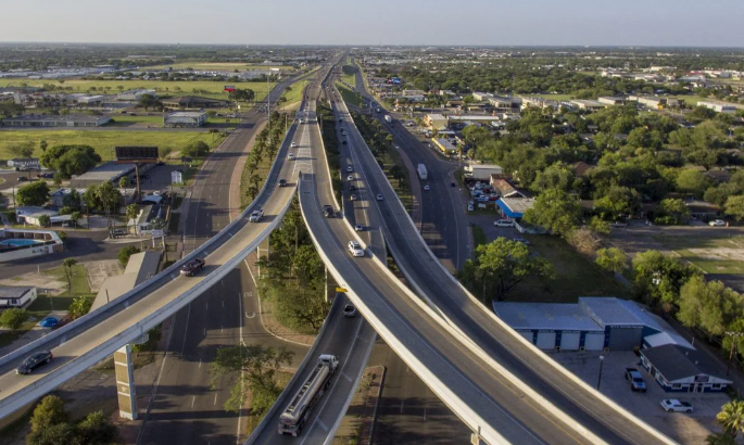 Snow-capped mountains rise behind a lake and evergreen trees under a blue sky with fluffy white clouds.