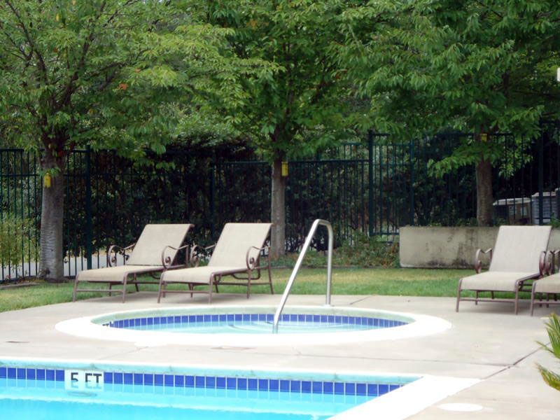 A pool with a jacuzzi, lounge chairs, and trees in a fenced area.